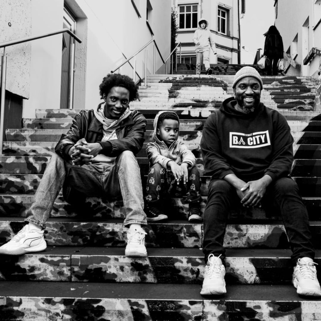 Zwei Erwachsene und ein Kind sitzen auf einer Treppe, das Foto ist in Schwarz-Weiß, sie sind leger gekleidet und schauen lächelnd in die Kamera.